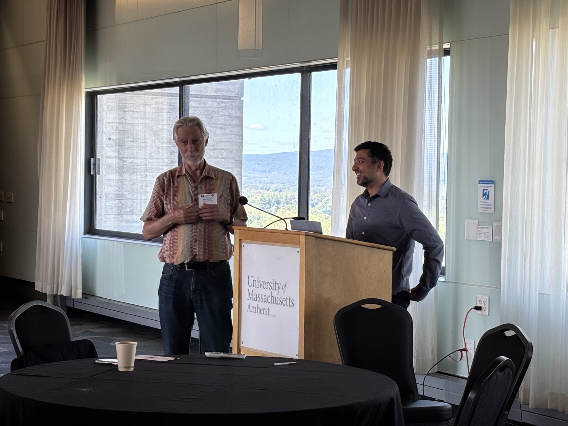 Two speakers stand beside the UMass Amherst podium at the NeSQom workshop.